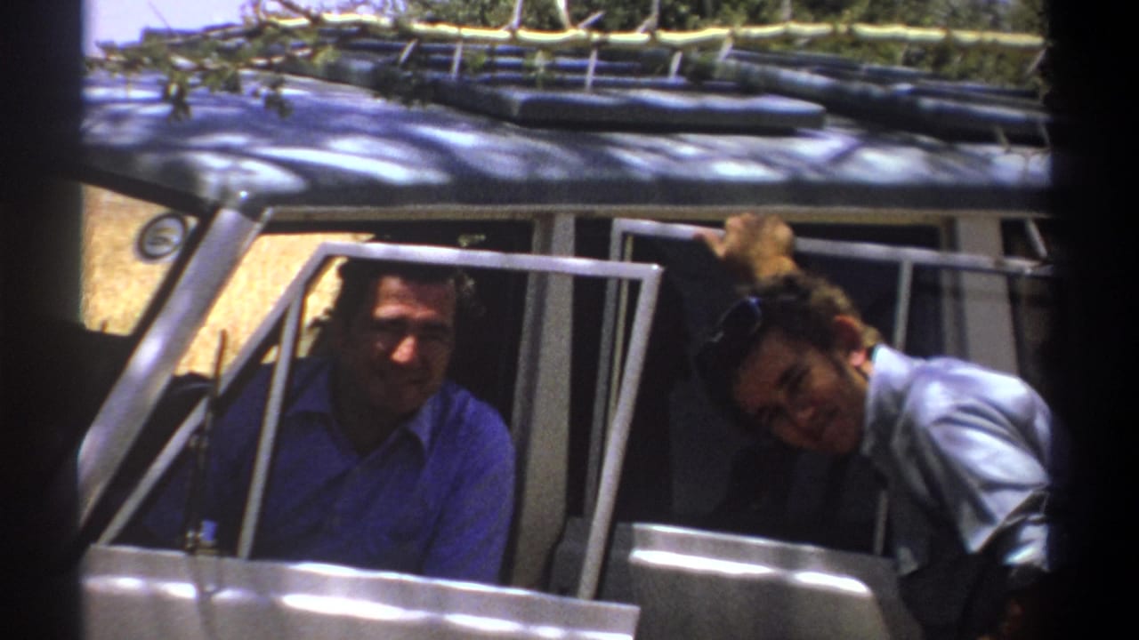 In Kenya 1969 home-movie footage, two adult men smile from a white safari-style vehicle with roof rack gear.
