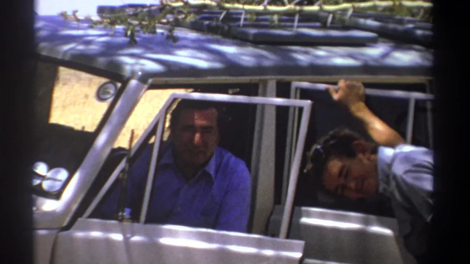The driver looks toward the camera as another man leans from the passenger side of a white vehicle in Kenya travel footage.