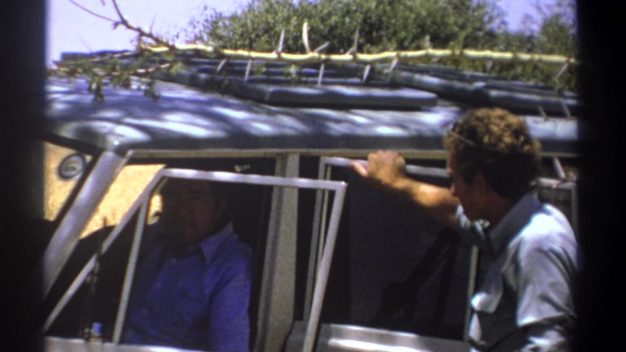 Man in grey shirt leans into an open vehicle while a man in blue sits inside.
