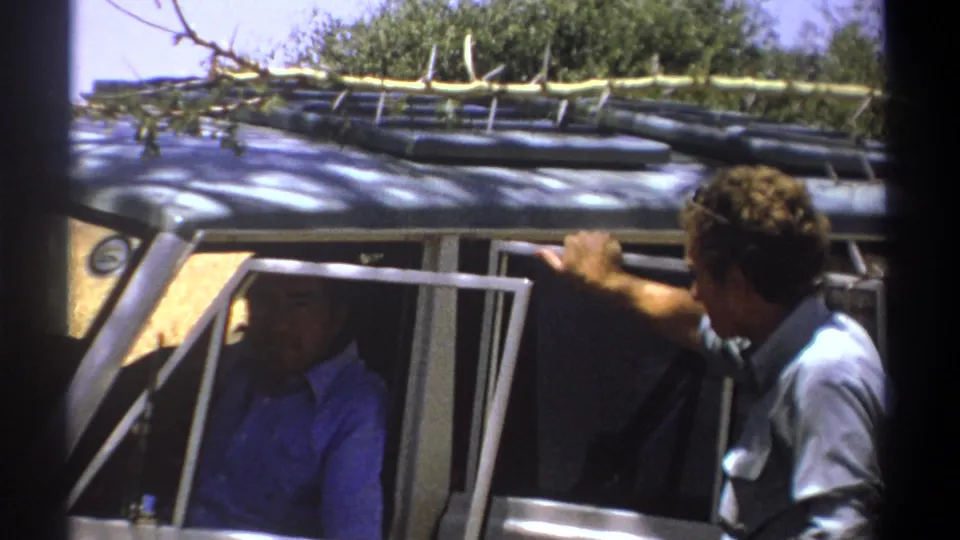 In 1969 Kenya footage, a man in a grey shirt leans at an open vehicle door as a man in blue sits in shadow inside.