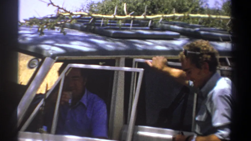 The grey-shirted man stands beside the open vehicle while the blue-shirted man remains seated inside in shadow.