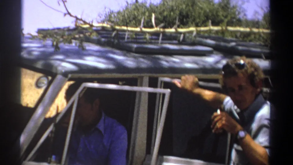 In a handheld 8mm frame from Kenya, the outside man shifts position near the open vehicle door as the seated man remains inside.