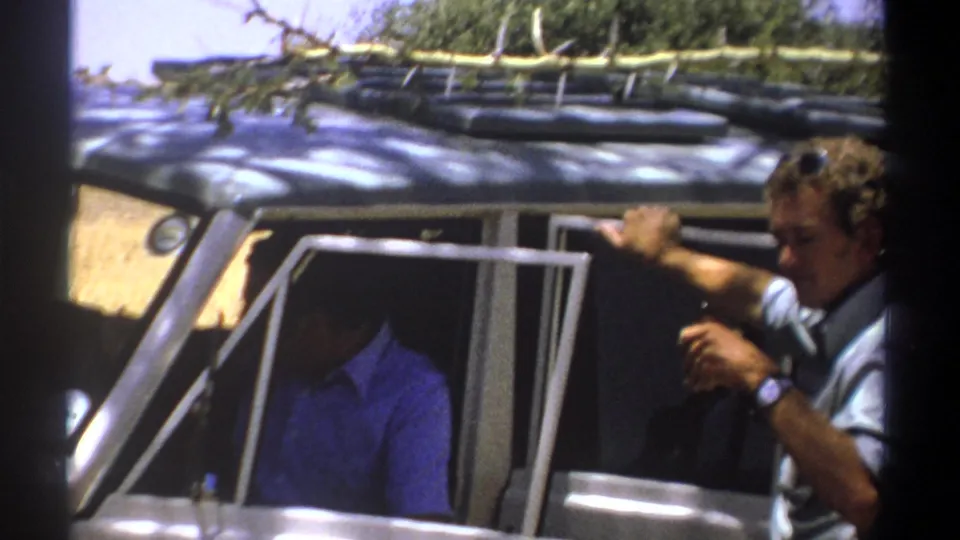 The grey-shirted man remains by the open vehicle in 1969 Kenya footage, with the blue-shirted passenger partly hidden by shadow.
