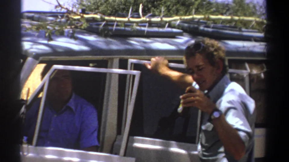 The grey-shirted man leans toward the open vehicle window, holding a small drink as the blue-shirted passenger remains visible in shadow.