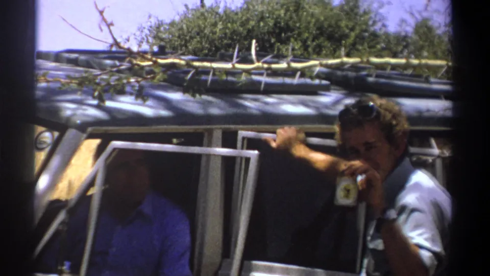 A man in a light grey shirt raises a drink near his face beside the vehicle, with the seated passenger still visible through the open window.