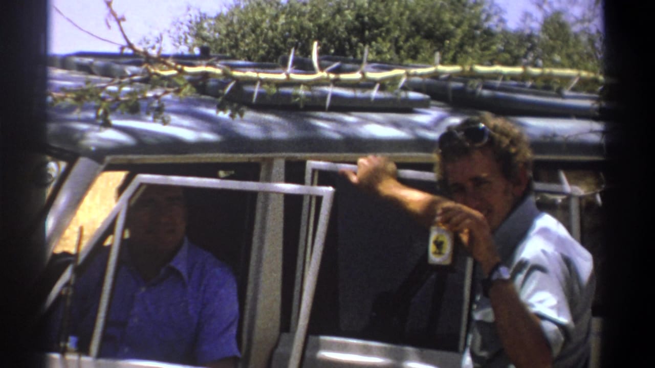 Two adult men in a safari vehicle, one holding a bottle near an open door.