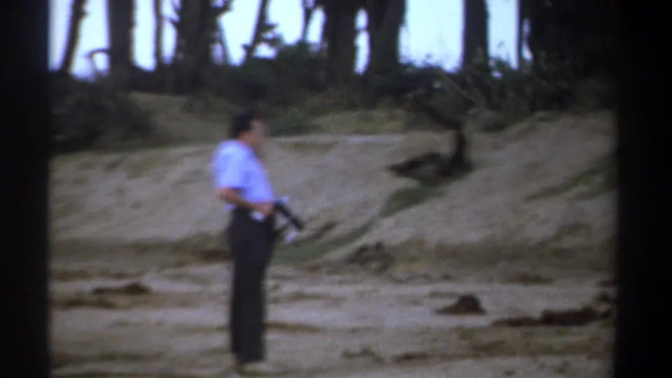 Kenya 1969 8mm frame showing a man in a light shirt and dark trousers holding a rifle in a sparse landscape.