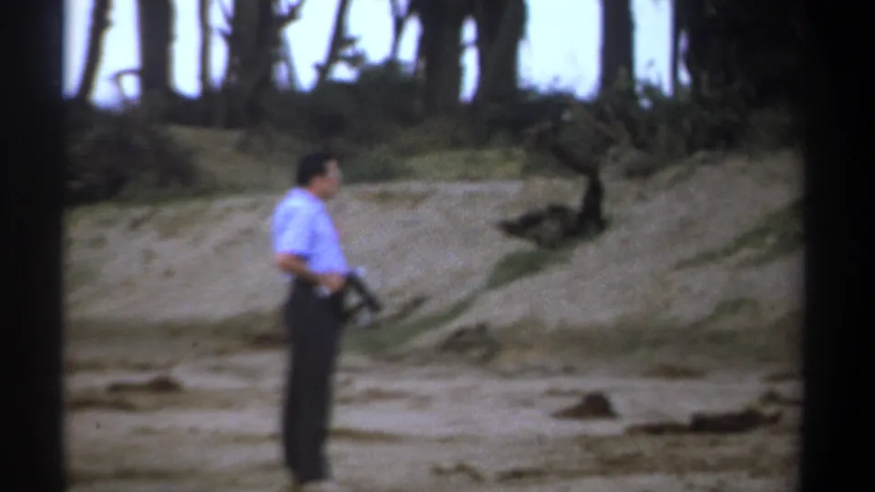 In Kenya 1969 safari footage, a man in a light blue shirt stands on a dusty roadside holding a rifle near scrub and trees.