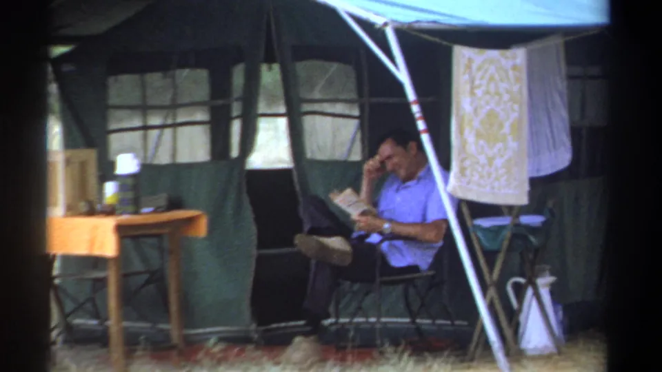 Adult man reads in a folding chair beside a green canvas safari tent and camp table in 1969 Kenya footage.