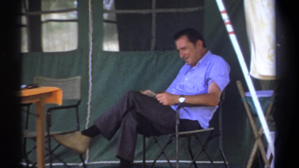 Adult man in a light blue shirt reads while seated by a green canvas tent in 1969 Kenya safari-camp footage.