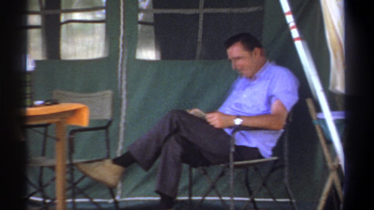 In Kenya 1969 home-movie footage, a man in a light blue shirt sits in a folding chair inside a green tent and reads.