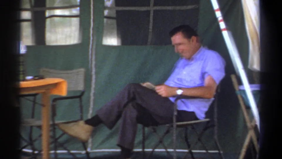 In Kenya 1969 home-movie footage, a man in a light blue shirt sits in a folding chair inside a green tent and reads.