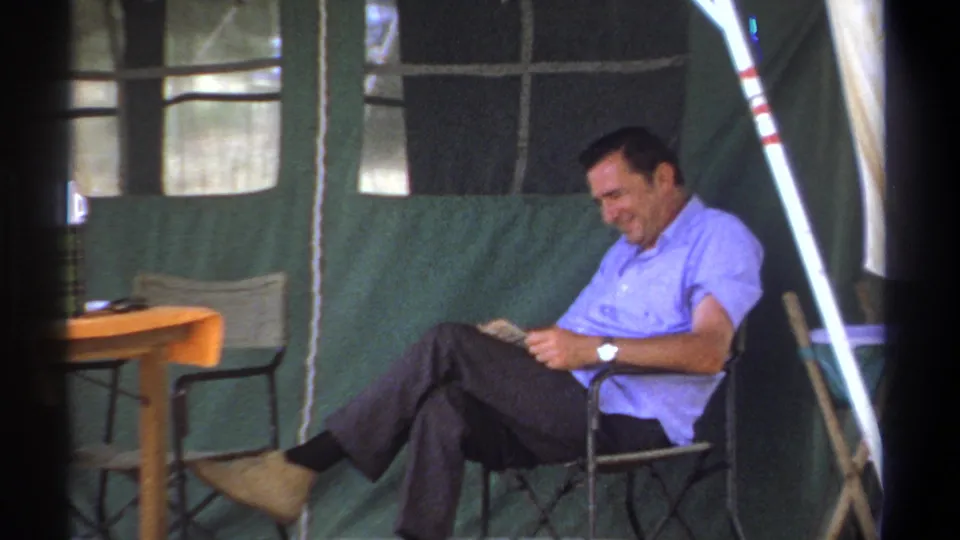 The man smiles while reading in a folding chair inside the green tent, with camp furniture visible around him.