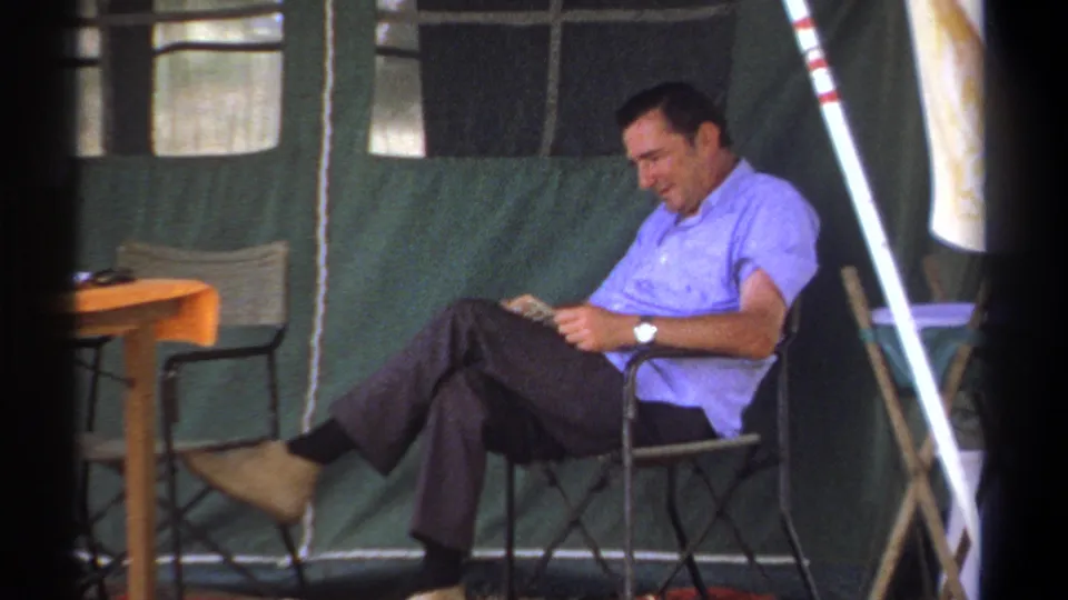 A relaxed seated man reads with crossed legs inside the Kenya camp tent, framed by soft 8mm film grain.