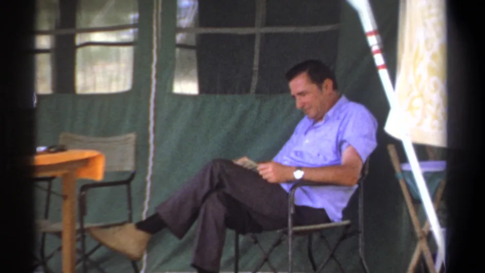 The man remains seated and reading inside the green canvas tent, with camp furniture and tent supports in view.