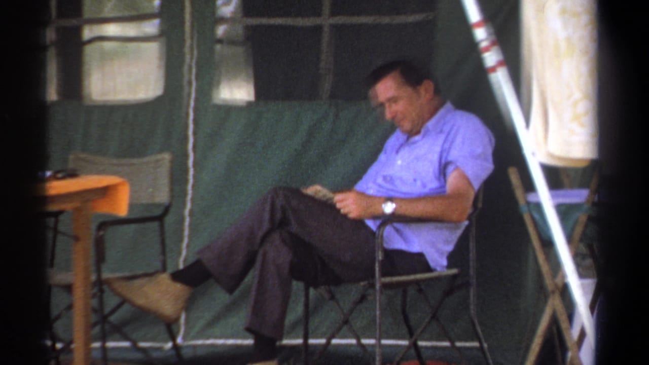 Man sitting in a folding chair inside a green canvas tent while reading.