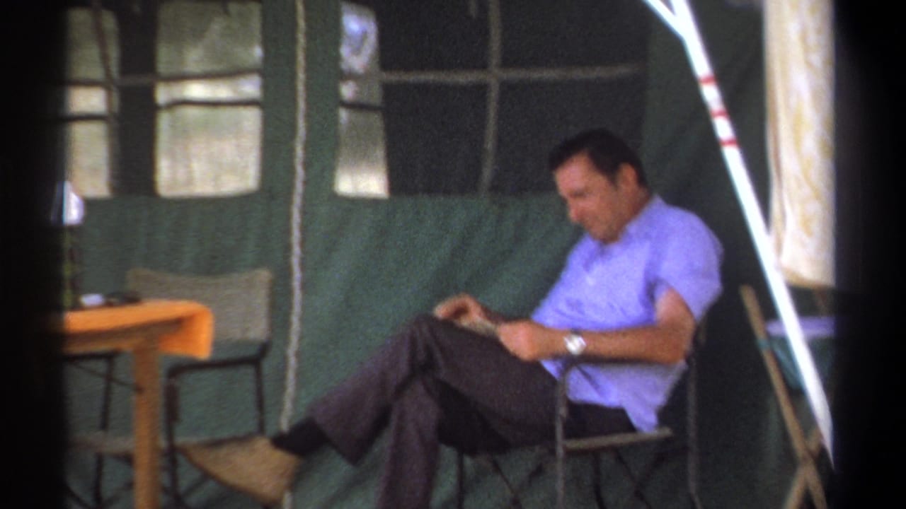 Seated man looks down at reading material beside a small camp table.