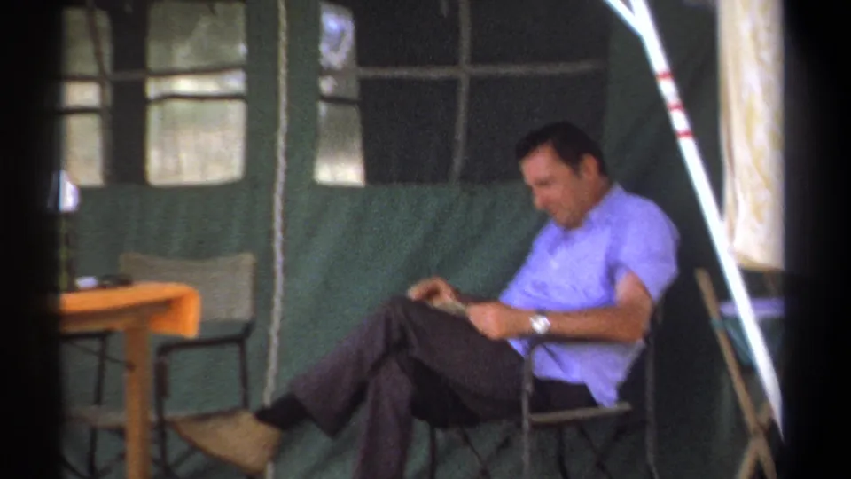 The 1969 Kenya home-movie frame shows a man reading in a canvas tent, with a small table and tent windows nearby.