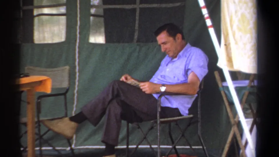 A man in a light blue shirt reads while seated in a folding chair inside a green canvas tent in Kenya, 1969.