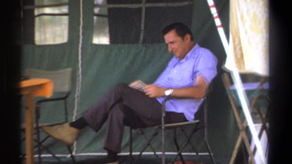 In this Kenya 1969 frame, a man sits cross-legged in a folding chair and reads inside a green canvas tented camp area.