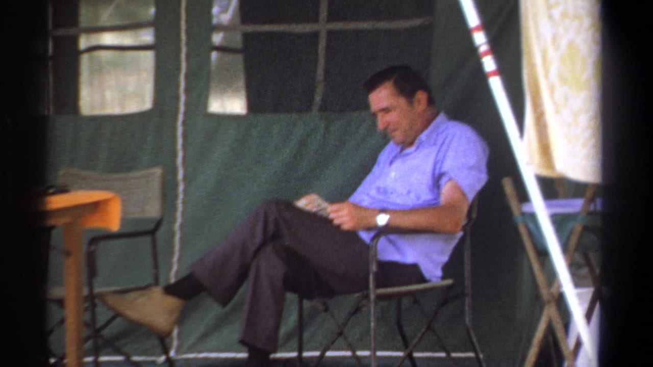Man in a blue short-sleeved shirt reads in a folding chair inside a green tent.
