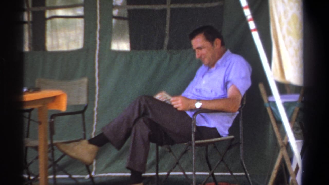 Man sits in a chair inside a tent, looking down at a book near a small table.