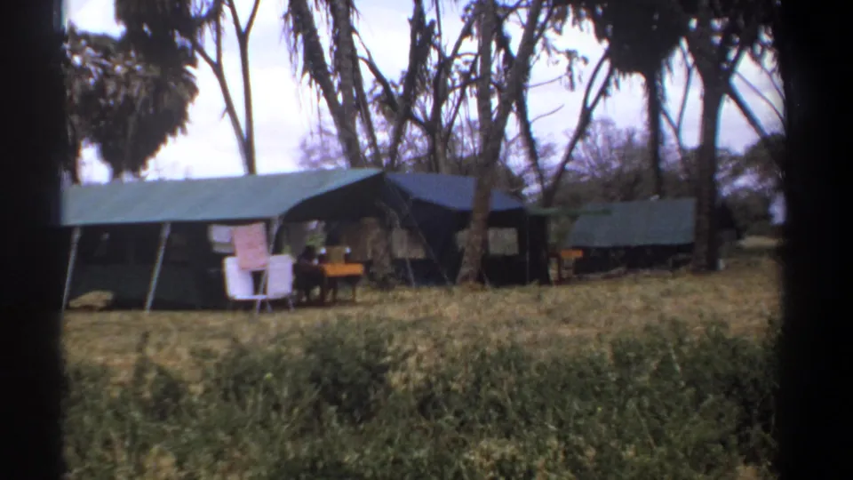 The final frame holds on the tented campsite, showing green canvas shelters, trees, dry ground, and camp furniture in Kenya.