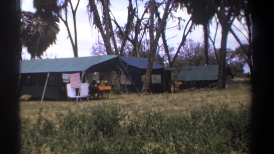1969 Kenya 8mm frame showing canvas safari tents under tall trees, with folding chairs, a small table, dry grass, and a pink towel.