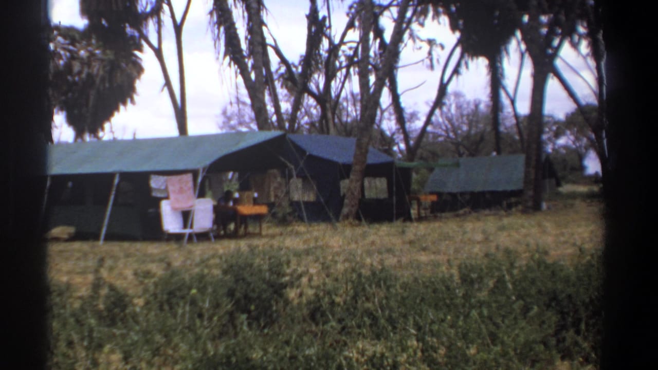 Dark canvas tents and outdoor seating sit beneath trees in a dry grassy camp area.