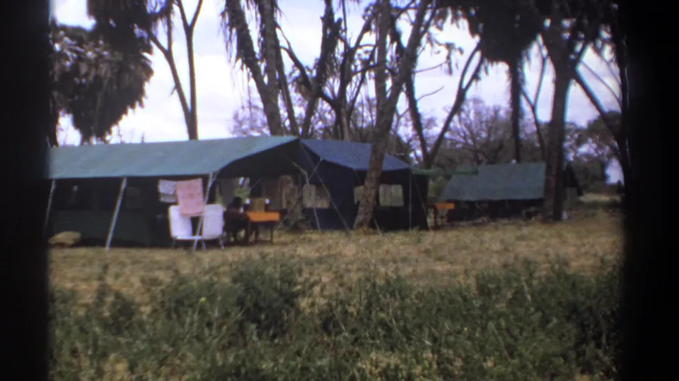1969 Kenya home-movie frame showing a row of safari tents beneath trees with camp furniture and a towel outside.