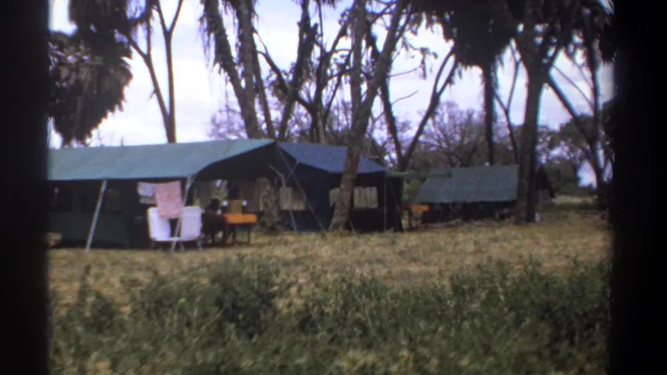 1969 Kenya 8mm frame of a safari camp with canvas tents, tall trees, outdoor seating, a small table, and dry grass.