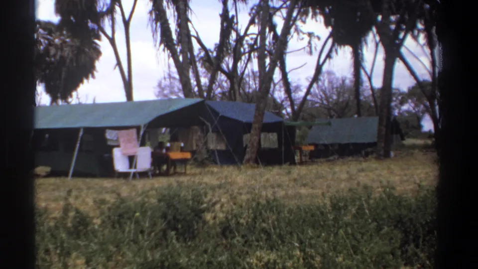 1969 Kenya home-movie view of green canvas tents beneath trees, with pale chairs, a table and dry grass filling the foreground.