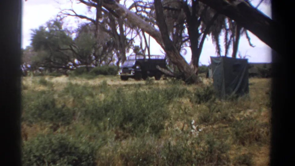 1969 Kenya frame showing a dark vehicle near canvas tents and trees, with dry grass and camp structures in the background.