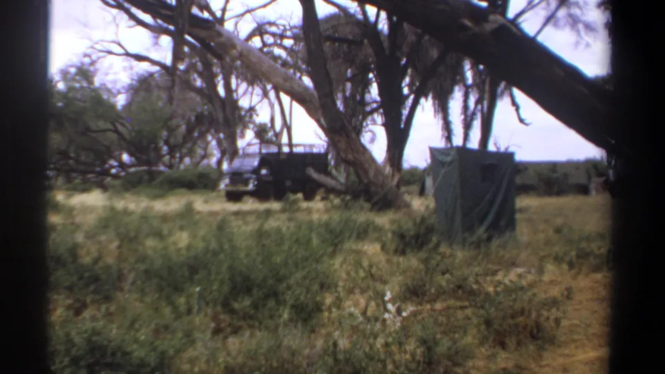 1969 Kenya home-movie frame showing tall grass, tree branches, a dark utility truck, and a small canvas tent at a bush camp.