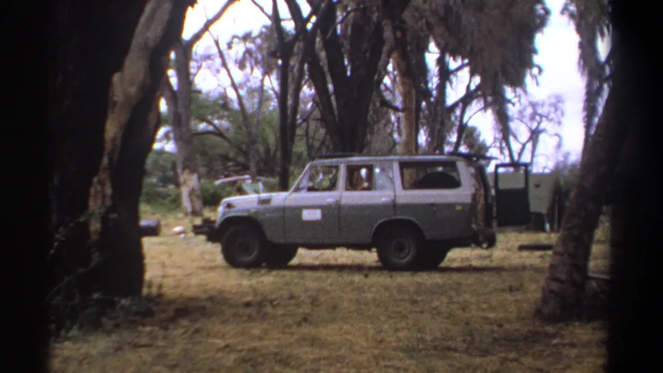 1969 Kenya home-movie frame showing a grey SUV in profile under trees, with a faint occupant visible through the side window.