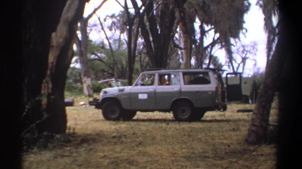 1969 Kenya frame of a grey vintage SUV parked among trees in a dry camp clearing, with the rear door open and a faint occupant visible.