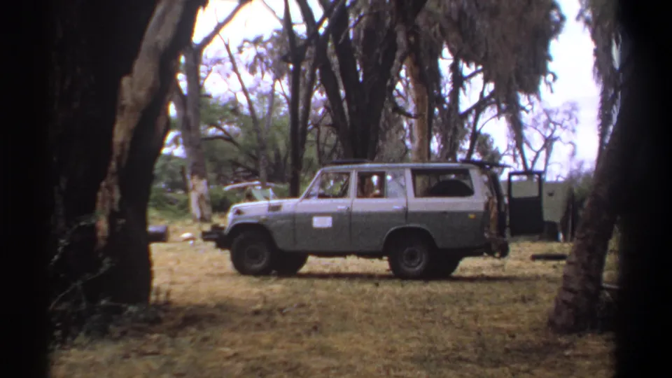 1969 Kenya home-movie frame with a parked two-tone station wagon in a wooded clearing and an occupant visible through the window.
