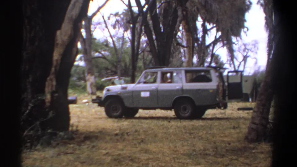 Final 1969 Kenya frame of a stationary two-tone utility vehicle in a wooded clearing, viewed through foreground trees.