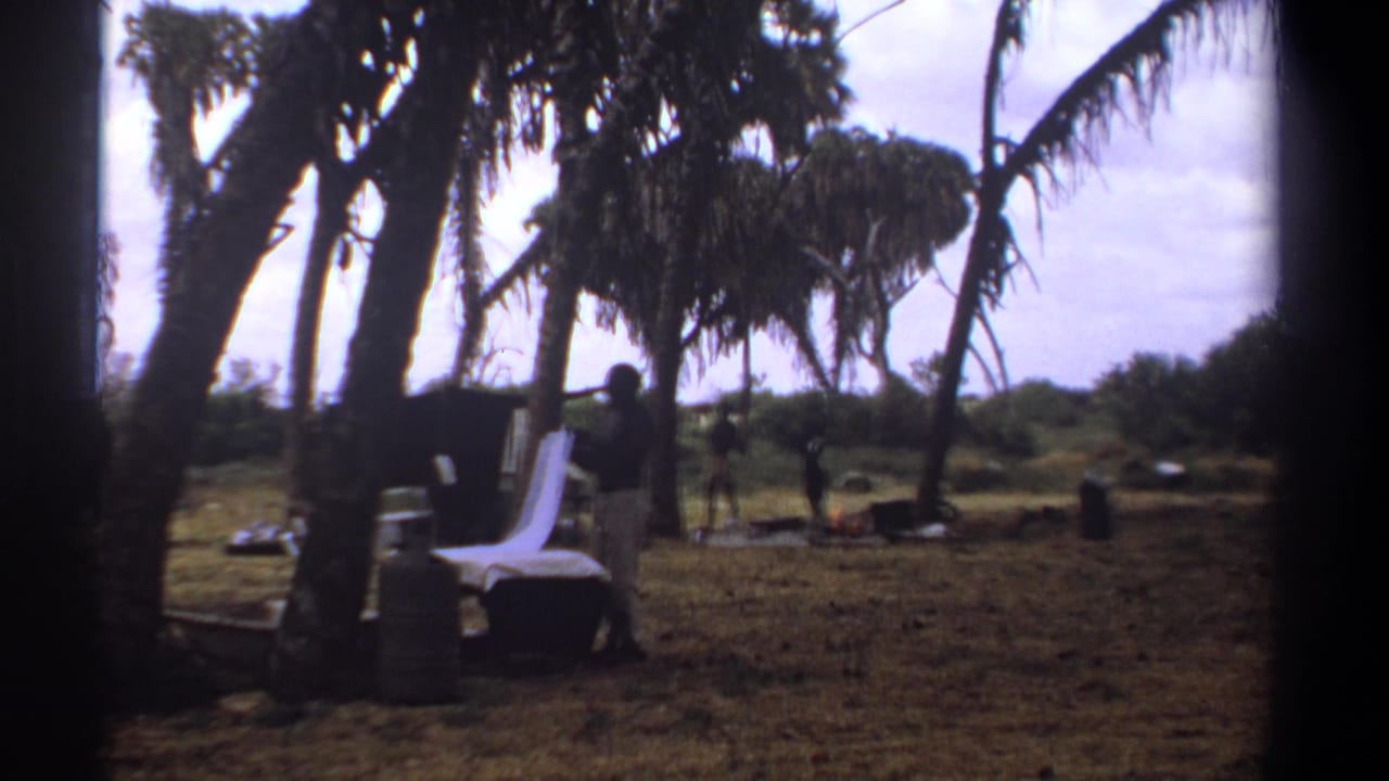 1969 Kenya home-movie frame showing men gathered beneath palm trees near a white-covered table or cot and scattered outdoor supplies.