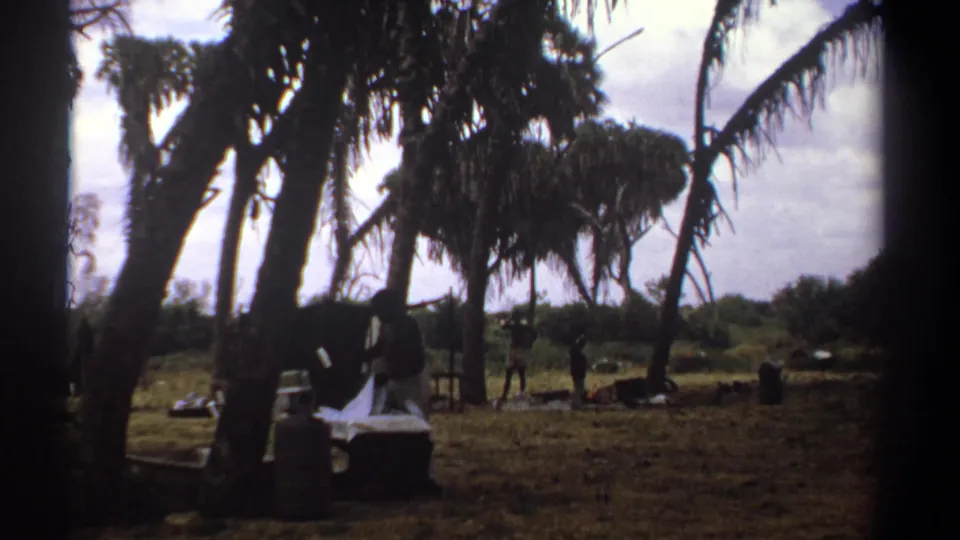 1969 Kenya 8mm frame with a person near a draped white sheet, containers, dry grass, and tall palm trees.