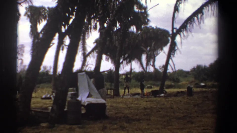 1969 Kenya 8mm frame of a white sheet draped near containers, with people and palm trees in the background.