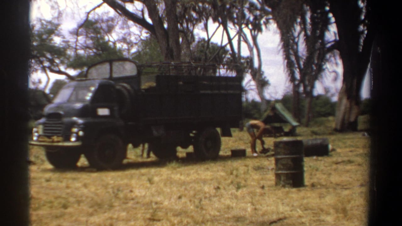 Two men work beside a black utility truck in a dry field with barrels and trees.