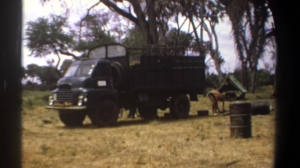 1969 Kenya 8mm frame of two men beside a black utility truck in a dry grassy field, with barrels, trees, and a small tent visible.