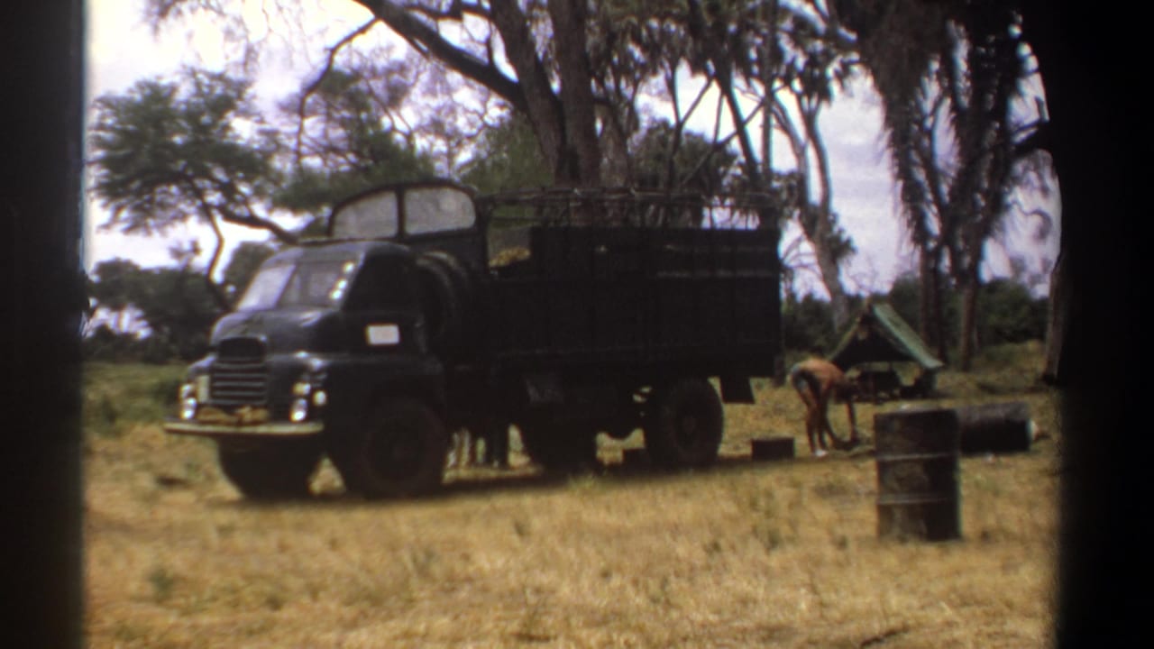 Black military-style truck parked near dry grass, trees, a small tent, and a person bending beside camp gear.