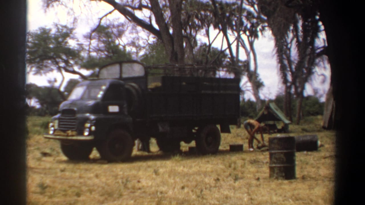 1969 Kenya 8mm frame showing a dark military-style truck at a dry savanna camp with barrels and scattered trees nearby.