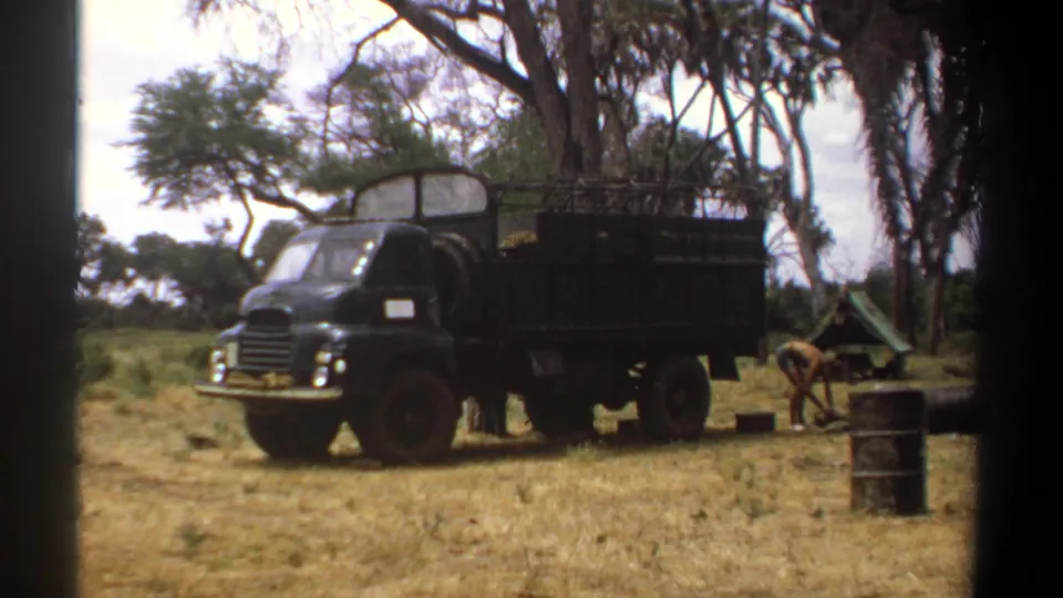 A dark military-style truck sits in a Kenyan savanna clearing, with a man working near barrels and field equipment.