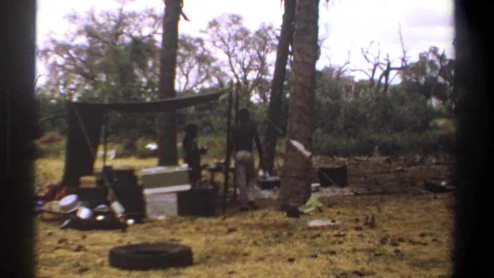 Two adult men are visible near a tarp-covered camp area in Kenya, with boxes, tires, trees, and dry savanna grass around them.