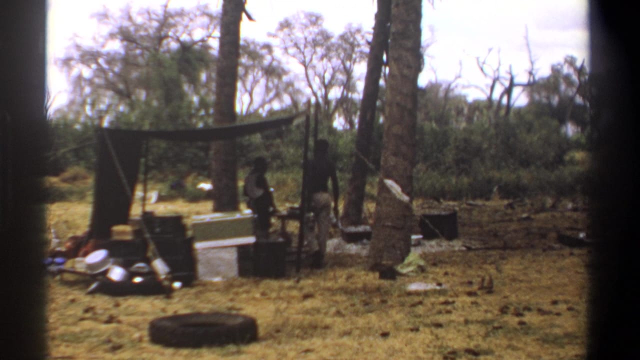 Two figures stand near a tarp shelter among trees with camp supplies on dry grass.