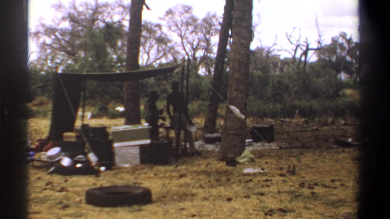A person moves beside a tarp shelter while camp gear sits under trees.