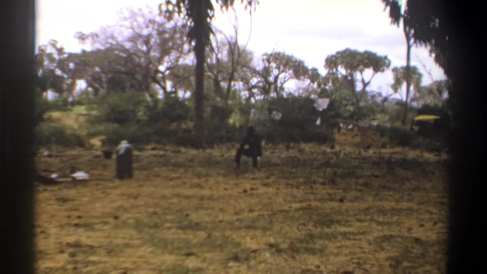 1969 Kenya home-movie view of a standing person, a seated figure, scattered items, dry grass, and wooded background.